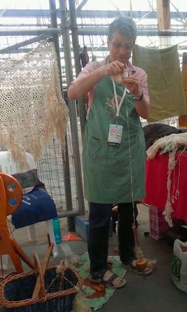 Suzy is standing up in front of her stall's display, holding her drop spindle by the yarn while the spindle spins near her ankles in front of her.