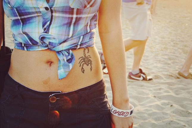 chica jovencita en la playa, con la camisa atada vemos un tatuaje en su hombligo, es el tatuaje de un escorpion
