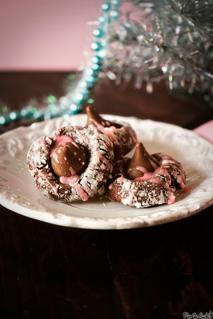 three chocolate thumbprint cookies on a white plate topped with Hershey kiss candies and a pink glaze with a silver Christmas tree in the background