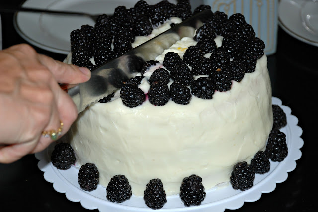 whole cake on a cake platter with white frosting and blackberries all around the bottom and blackberries all over the top cutting into it with a big knife 