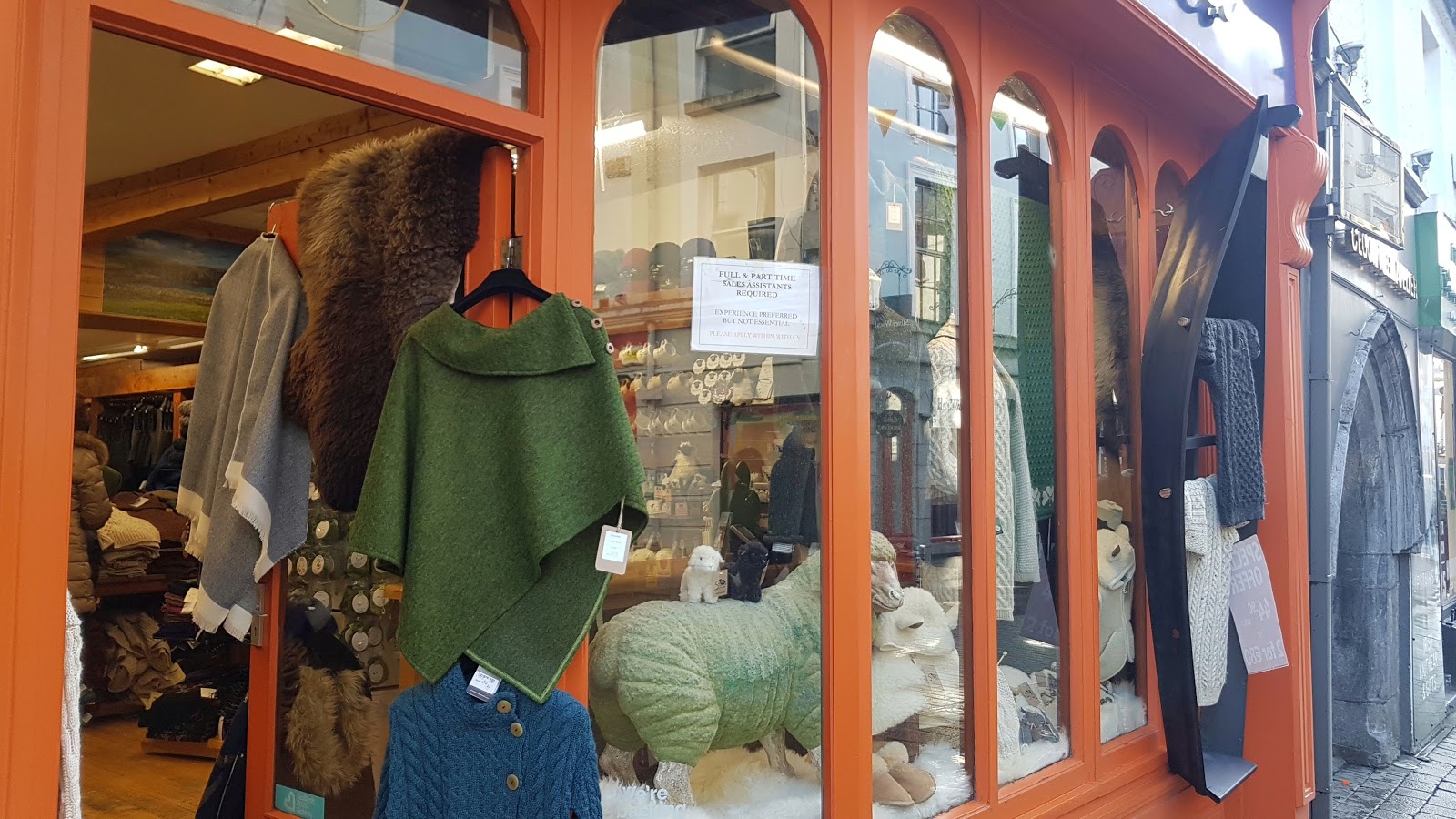 shop-front with job advertisement sign, and green knitted poncho on a hanger, along with a sheepskin rug and a grey knitted jumper