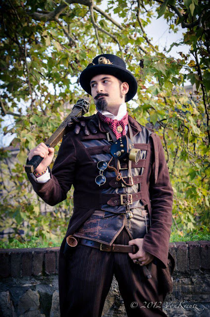 Man wearing steampunk clothing (top hat, goggles, coat, waistcoat, trousers) carrying a wooden club wrapped in barbed wire
