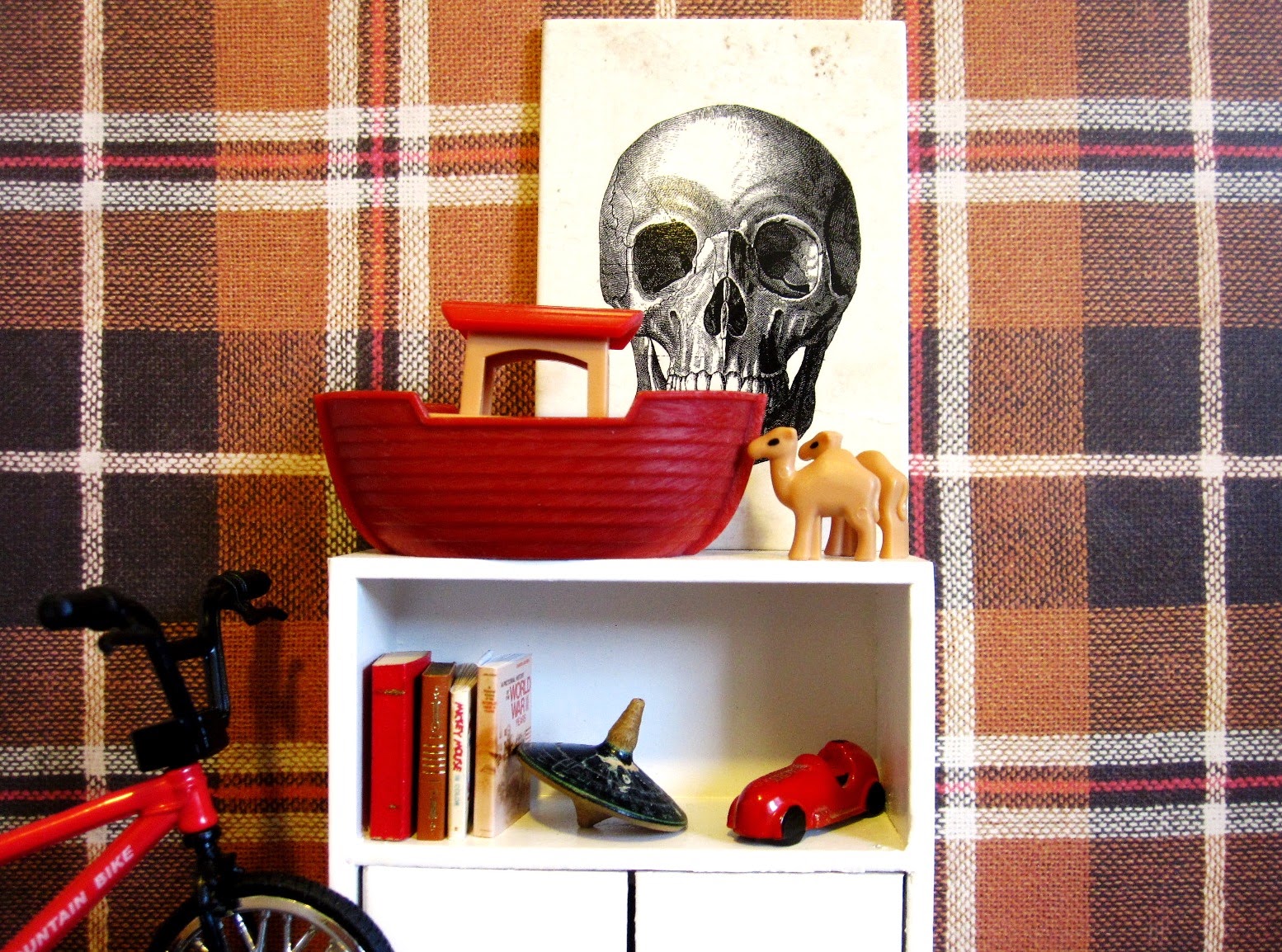 Modern dolls' house scene of part of a boy's room, with fawn, red and white tartan wallpaper and a white shelf holding a Noah's ark, a toy car, some books and a spinning top. To one side is a BMX-style bike.