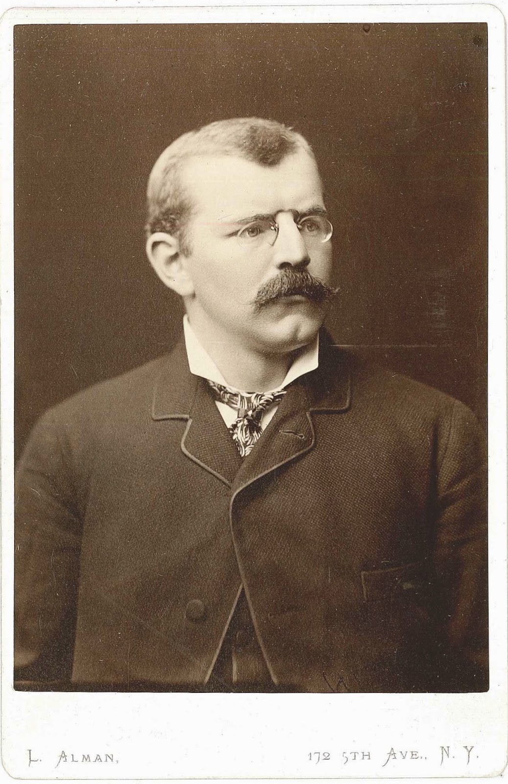 A black and white photograph of a man with a mustache and glasses.