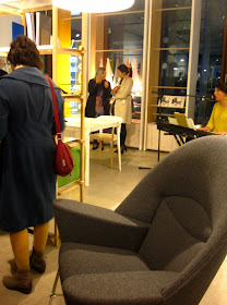 An exhibition space with an armchair and table and chair on display. A musician sings and plays keyboard in the corner.