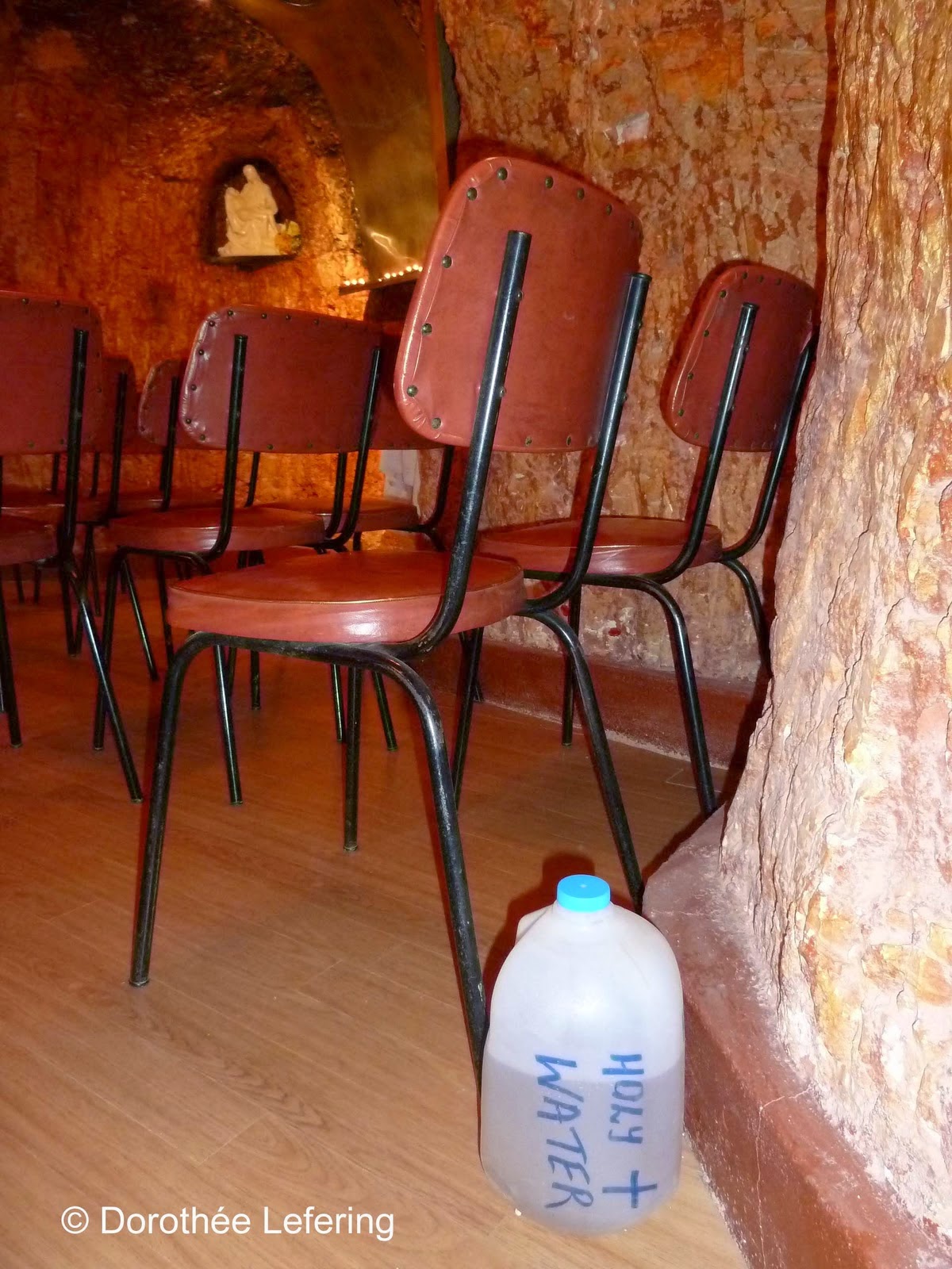 A plastic canister labelled with Holy Water, next to brown chairs with a figure of a madonna and the stone walls of the church as a background