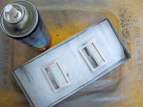 Box lid containing undercoated dolls' house miniature windows, sitting next to a can of spraypaint on the lid of a recycling bin.