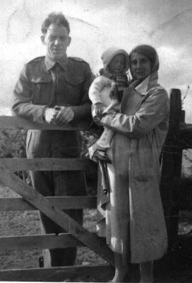 A man in army uniform leaning on a five barred gate, on this side separated from him by the gate is a woman wearing a head scarf and long over coat holding a small child