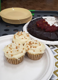 Plate of cupcakes next to a plate holding half a dark chocolate cake with fresh raspberries on the top of it. Next to the cake is a stack of plates, and in front is a stack of paper napkins.