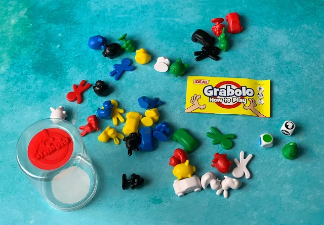 Different shape plastic pieces in white, yellow, red, green, blue, black and yellow and 2 dice next to instructions for How to Play Grabolo
