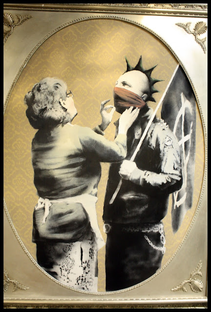 Untitled work by Banksy from his Bristol Museum Vs. Banksy show. The image depicts an anarchist, with a spiky mohawk, red scarf tied around his nose and mouth, and a black flag on a pole held against his shoulder. In front of him stands his mother, who is trying to straighten his red scarf before he goes out. Photograph by Josh Blair.