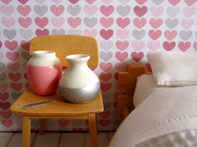 Modern dolls' house miniature bedroom scene with futon bed with a white waffle-weave blanket, a blonde-wood chair holding two dip-painted white vases and a feather.