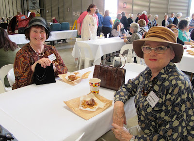 Two women dressed in 1940s outfits, sitting on plastic chairs and drinking coffee and eating muffins.