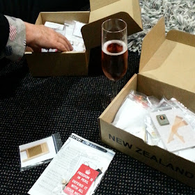 Two open cardboard boxes in the style of Red Cross boxes. Between them is a glass of sparkling wine, and a hand reaches into the back one.