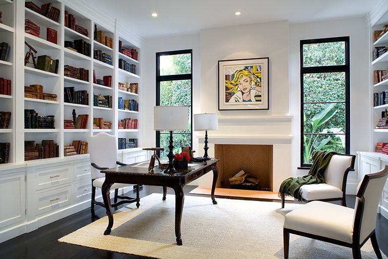 Home office in a Spanish revival home with white built in bookshelf, traditional wood desk and wood chairs with white seats and backs, a fireplace with a a Roy Lichtenstein print on the mantel and black paned windows