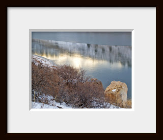 A framed photo of a hillside covered in Mountain Mahogany drops steeply to an ice covered lake reflecting the setting sun.