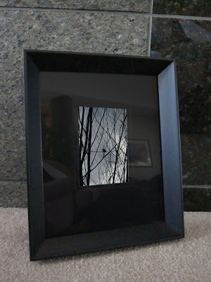 A black matted and framed photo of a small bird silhouette resting on tree branches against a gray storm clouded sky.