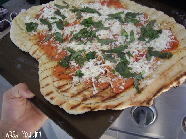 A hand holding a cookie sheet with grilled pizza crust, tomatoes, shredded cheese and chopped spinach. 