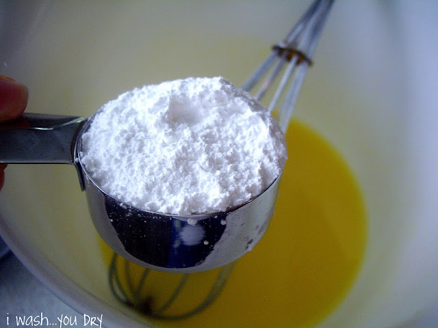 A measuring cup of powdered sugar above a bowl of yellow liquid. 