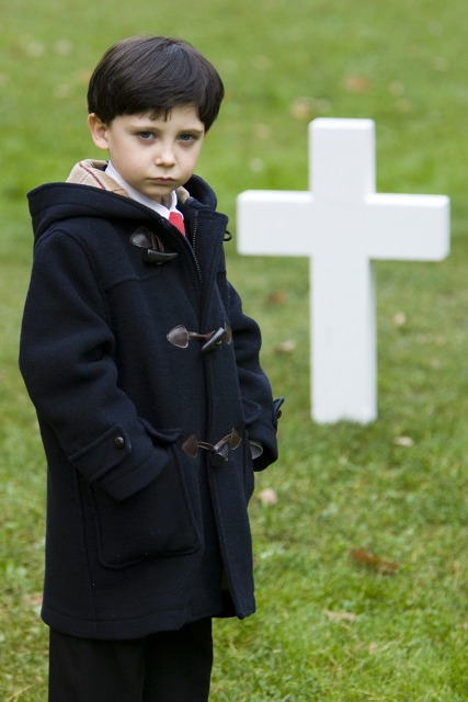 Damien em pé em um cemitério (um gramado e uma cruz lisa branca quase da altura do menino) usando casaco preto grosso e longo, com as mãos no bolso.