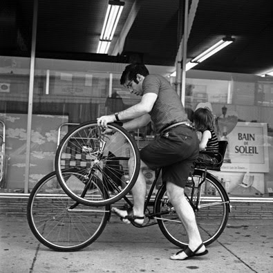 60s photo of a young man mounting a bike with an extra wheel in his hand, young child sitting in seat on back