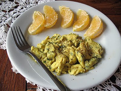 A close up photo of scrambled eggs with garlic, basil and feta with mandarin orange slices to to side on a white plate.
