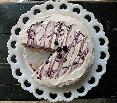 An overhead photo of a vanilla bean mulberry cake on a white cake stand topped with fresh mulberries and a slice missing.